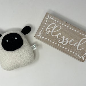 Pode incluir: Um brinquedo de pel&uacute;cia de ovelha branco e felpudo com orelhas, olhos e nariz pretos. O brinquedo est&aacute; sentado em uma superf&iacute;cie branca ao lado de uma placa de madeira que diz "blessed" em letras brancas.
