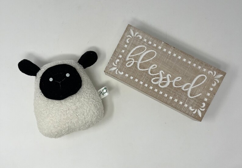 Pode incluir: Um brinquedo de pel&uacute;cia de ovelha branco e felpudo com orelhas, olhos e nariz pretos. O brinquedo est&aacute; sentado em uma superf&iacute;cie branca ao lado de uma placa de madeira que diz "blessed" em letras brancas.