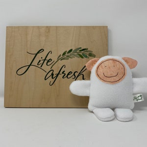 May include: White plush toy shaped like a sheep with a smiling face. The toy has a brown felt face with a stitched smile and a tag that says "Life Afresh".