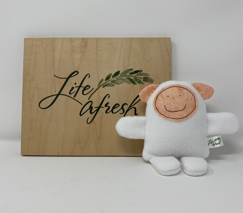 May include: White plush toy shaped like a sheep with a smiling face. The toy has a brown felt face with a stitched smile and a tag that says "Life Afresh".