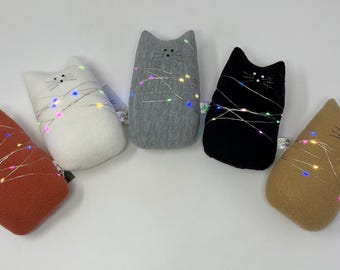 May include: Five cat-shaped plush toys in rust, white, grey, black, and tan. Each cat has embroidered facial features and is wrapped with a string of colourful LED lights. A whimsical and decorative item.