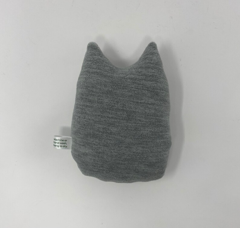 May include: Gray plush cat-shaped pillow with a white tag that reads "Machine or hand wash, hang to dry."