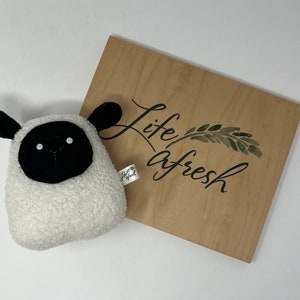 Pode incluir: Um brinquedo de pel&uacute;cia de ovelha branco e preto com uma cara preta e uma pele branca. O brinquedo est&aacute; sentado em uma placa de madeira que diz "life afresh" com um design de folha verde.