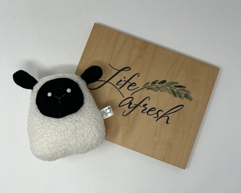Pode incluir: Um brinquedo de pel&uacute;cia de ovelha branco e preto com uma cara preta e uma pele branca. O brinquedo est&aacute; sentado em uma placa de madeira que diz "life afresh" com um design de folha verde.