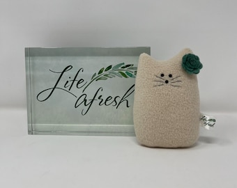 May include: A plush, beige cat-shaped toy with embroidered whiskers and a green flower on its head. The cat toy has a small tag attached. Beside it is a clear block with the words "Life a fresh" in script.