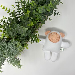 May include: White plush toy shaped like a sheep with a brown face and a stitched-on smile. The toy is lying on a white surface with a potted plant in the background.