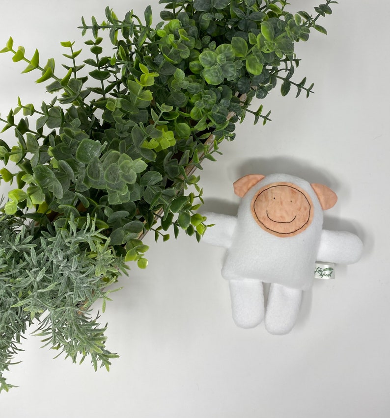 May include: White plush toy shaped like a sheep with a brown face and a stitched-on smile. The toy is lying on a white surface with a potted plant in the background.