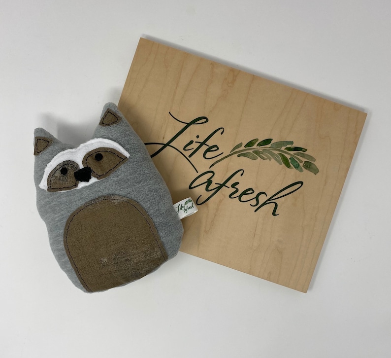 May include: A gray and brown raccoon plush toy with black eyes and a white nose. The toy is sitting on a wooden surface with the text "life afresh" and a green leaf design.