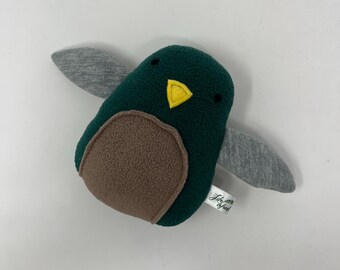 Puede incluir: Un juguete de pájaro de peluche hecho a mano en vuelo. El pájaro tiene un cuerpo verde oscuro, un pecho marrón, alas grises y un pico amarillo. El juguete tiene pequeños ojos negros y una etiqueta que dice "Life Seed".