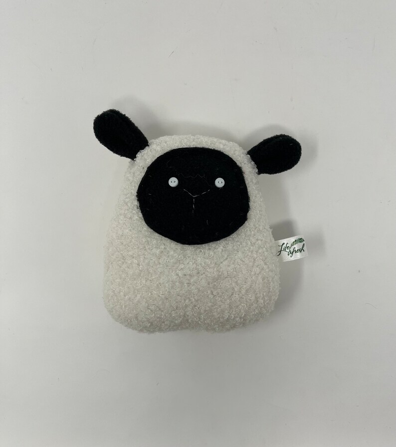 Pode incluir: Brinquedo de pel&uacute;cia de ovelha branca com orelhas, rosto e olhos pretos. O brinquedo tem uma pequena etiqueta verde com o texto "Little Sheep" nela.