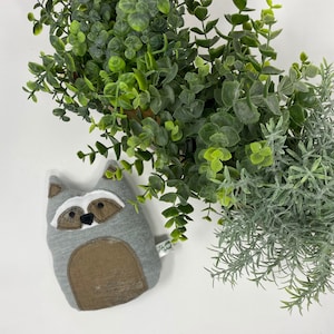 May include: A gray and brown plush raccoon toy with a white face and black eyes. The toy is lying on a white surface.