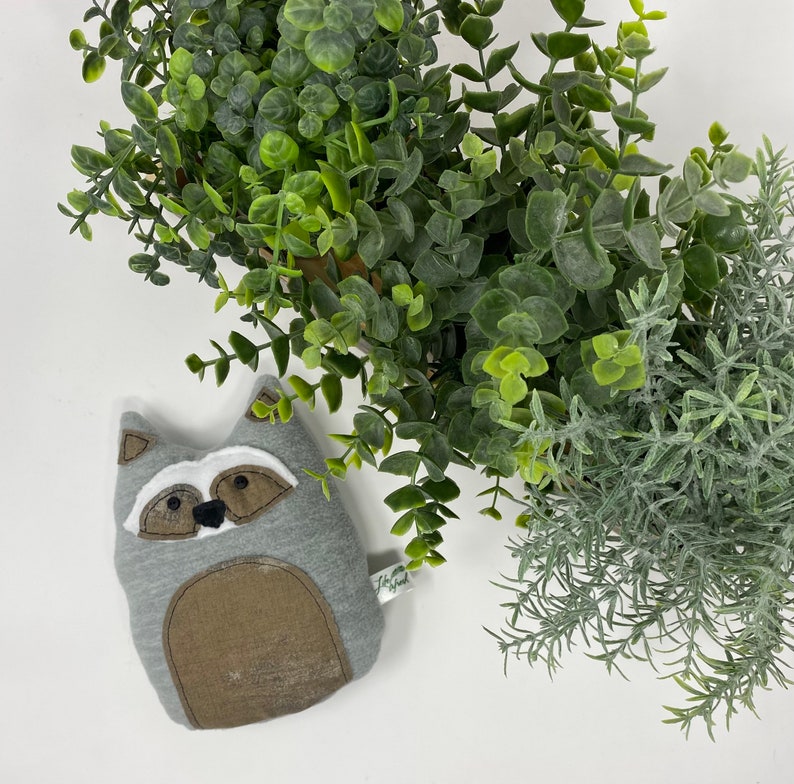 May include: A gray and brown plush raccoon toy with a white face and black eyes. The toy is lying on a white surface.