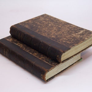 May include: Two antique books with brown leather covers and worn spines. The books are stacked on top of each other.