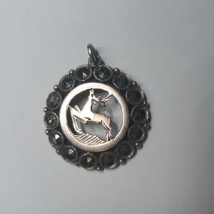 Puede incluir: Un colgante de plata con un diseño circular que presenta una silueta de ciervo recortada rodeada de piedras preciosas negras.