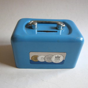 Può includere: Un salvadanaio di metallo blu chiaro con uno slot in cima e una maniglia. La parte anteriore del salvadanaio ha un adesivo con immagini di monete.