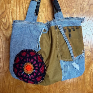 May include: A handmade tote bag constructed from repurposed denim and fabric scraps. The bag features a large, circular, crocheted patch with red, black, and orange details. The handles are made from denim and patterned fabric.