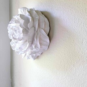 Puede incluir: Una escultura de pared de cer&aacute;mica blanca de una gran flor estilizada con m&uacute;ltiples capas de p&eacute;talos.