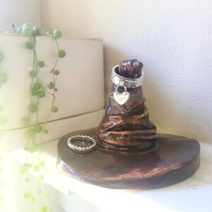 May include: A brown ceramic ring holder shaped like a wizard's hat. A silver ring with a heart-shaped charm and a second ring are displayed on the holder. The holder sits on a white surface.