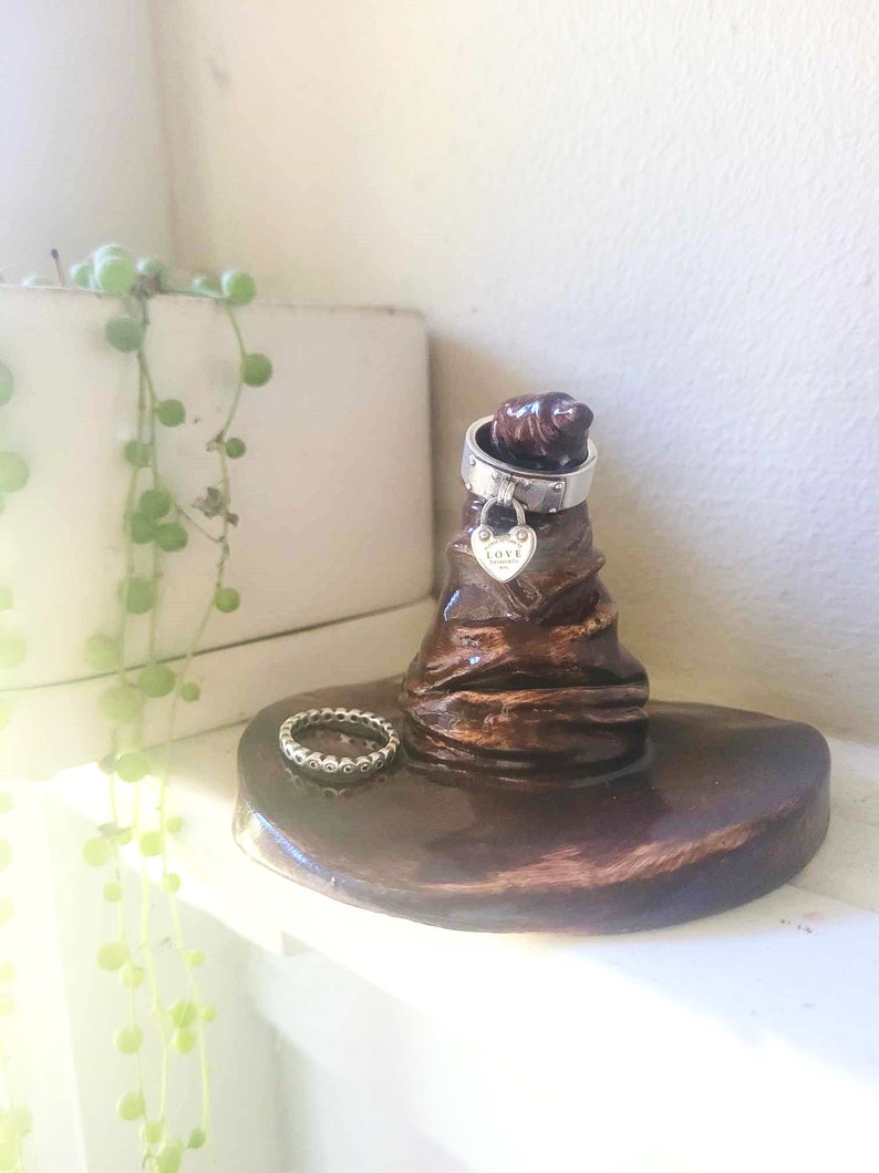 May include: A brown ceramic ring holder shaped like a wizard's hat. A silver ring with a heart-shaped charm and a second ring are displayed on the holder. The holder sits on a white surface.