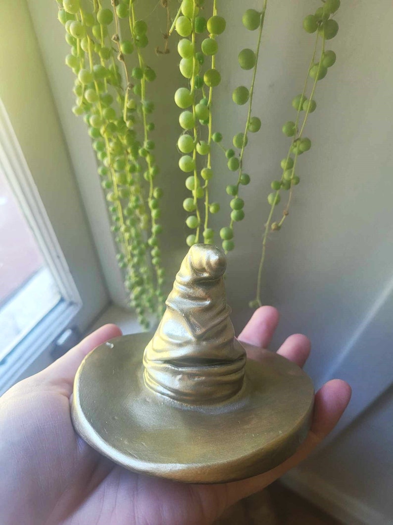 May include: A gold-colored, decorative hat with a wide brim. The hat is a replica of the Sorting Hat from the Harry Potter series. The hat is held in a person's hand, with a trailing plant in the background.