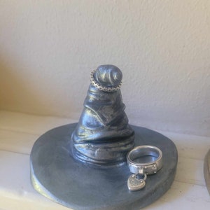 May include: A gray, handcrafted ring holder shaped like a witch's hat. Two silver rings are displayed: one on the hat's tip and another on the base. The base is round and painted gray. The ring on the base has a heart-shaped charm.
