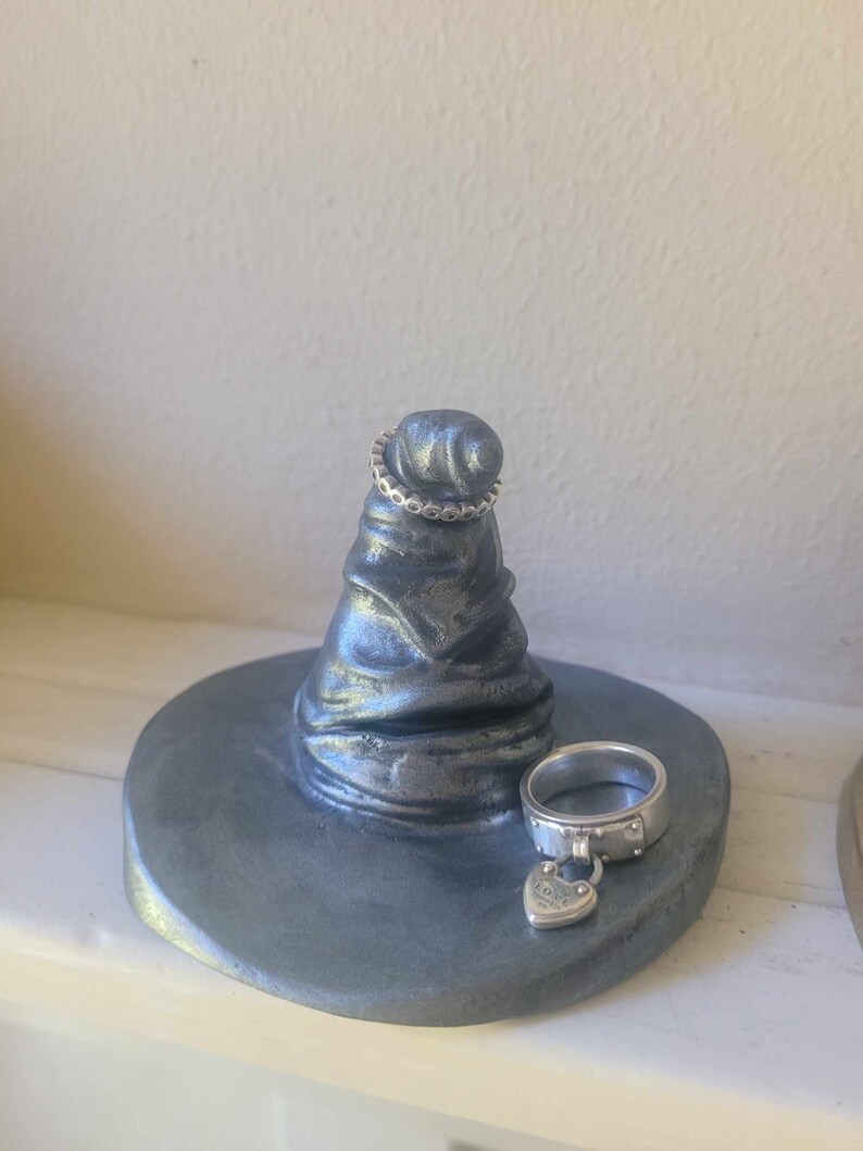 May include: A gray, handcrafted ring holder shaped like a witch's hat. Two silver rings are displayed: one on the hat's tip and another on the base. The base is round and painted gray. The ring on the base has a heart-shaped charm.