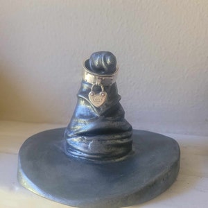 May include: A silver-colored ring holder shaped like a wizard's hat. A silver ring with a heart-shaped charm that says "LOVE" is on top of the hat. The hat is on a round base.