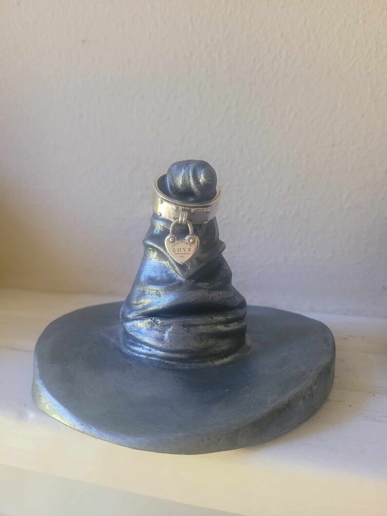 May include: A silver-colored ring holder shaped like a wizard's hat. A silver ring with a heart-shaped charm that says "LOVE" is on top of the hat. The hat is on a round base.