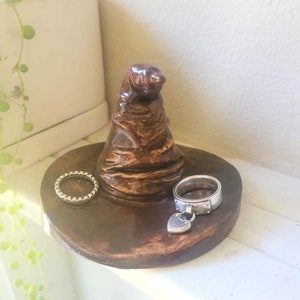 May include: A brown ceramic ring holder shaped like the Sorting Hat from the Harry Potter series. Two silver rings rest on the holder's base. The holder sits on a white surface, with a plant in the background.