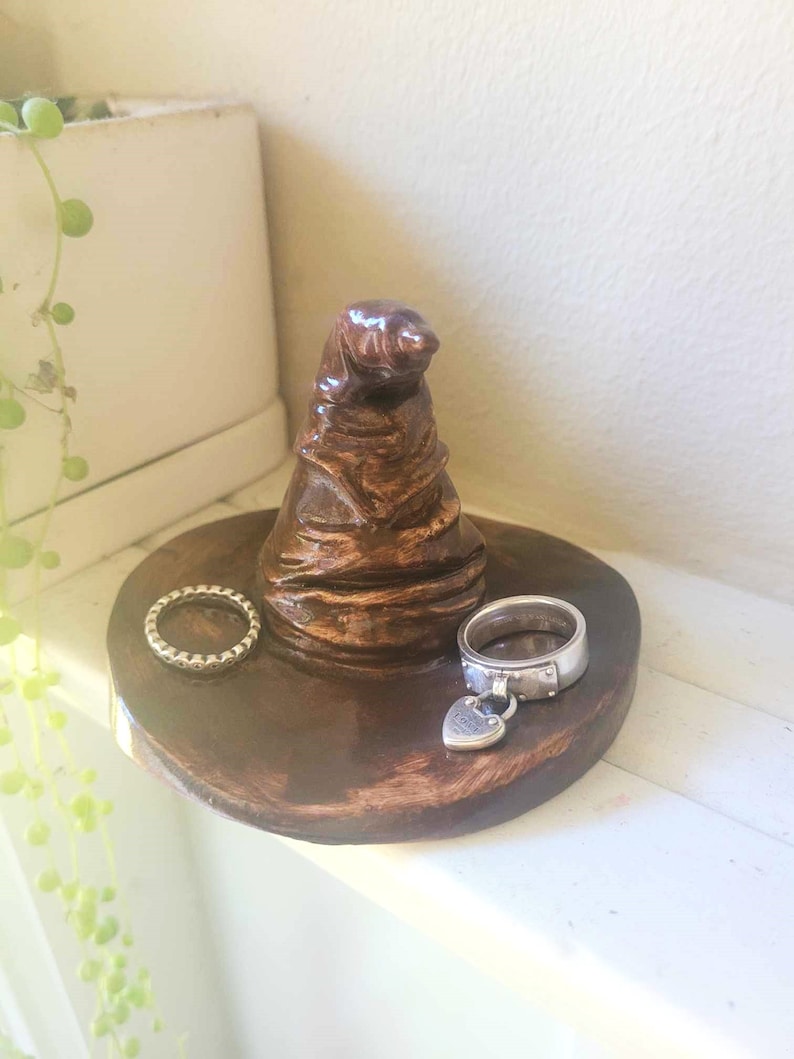 May include: A brown ceramic ring holder shaped like the Sorting Hat from the Harry Potter series. Two silver rings rest on the holder's base. The holder sits on a white surface, with a plant in the background.