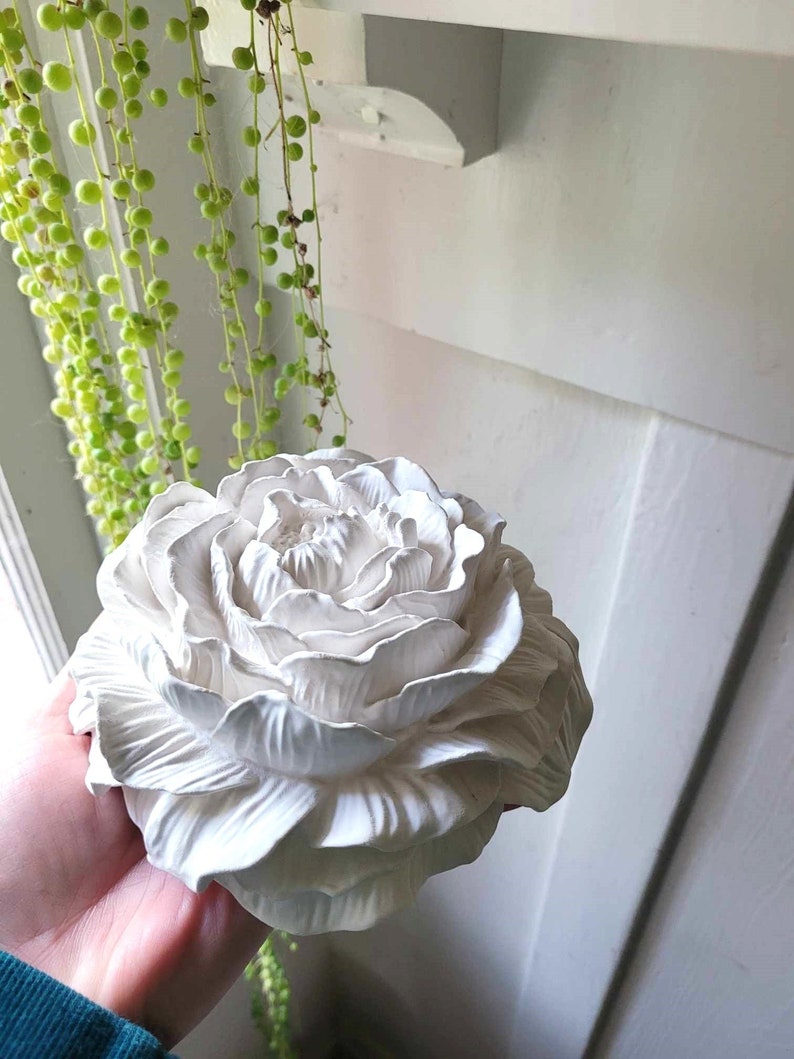 Puede incluir: Una flor de yeso blanco con un dise&ntilde;o detallado y realista. La flor tiene varias capas de p&eacute;talos y un centro que se asemeja a un capullo.