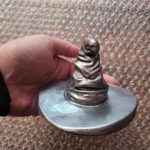 May include: A silver-colored, detailed replica of the Sorting Hat from the Harry Potter series. The hat has a wide brim and a textured, conical crown with folds and creases. The hat is held in a hand.