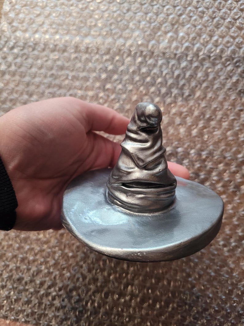 May include: A silver-colored, detailed replica of the Sorting Hat from the Harry Potter series. The hat has a wide brim and a textured, conical crown with folds and creases. The hat is held in a hand.