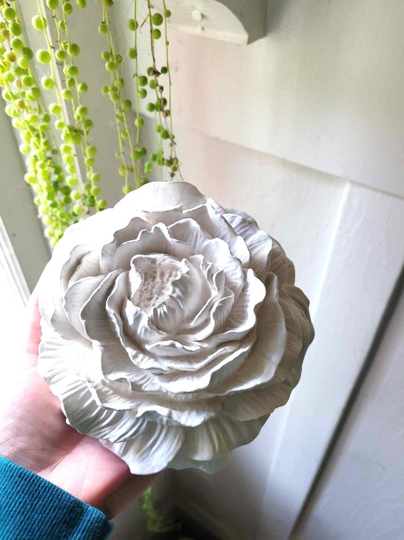 Puede incluir: Una gran flor de cer&aacute;mica blanca con un dise&ntilde;o detallado y en capas. La flor se sostiene en una mano, y el fondo es una pared blanca.
