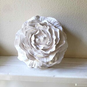 Puede incluir: Una flor de cer&aacute;mica blanca con un dise&ntilde;o detallado y en capas. La flor es una sola flor con m&uacute;ltiples p&eacute;talos.