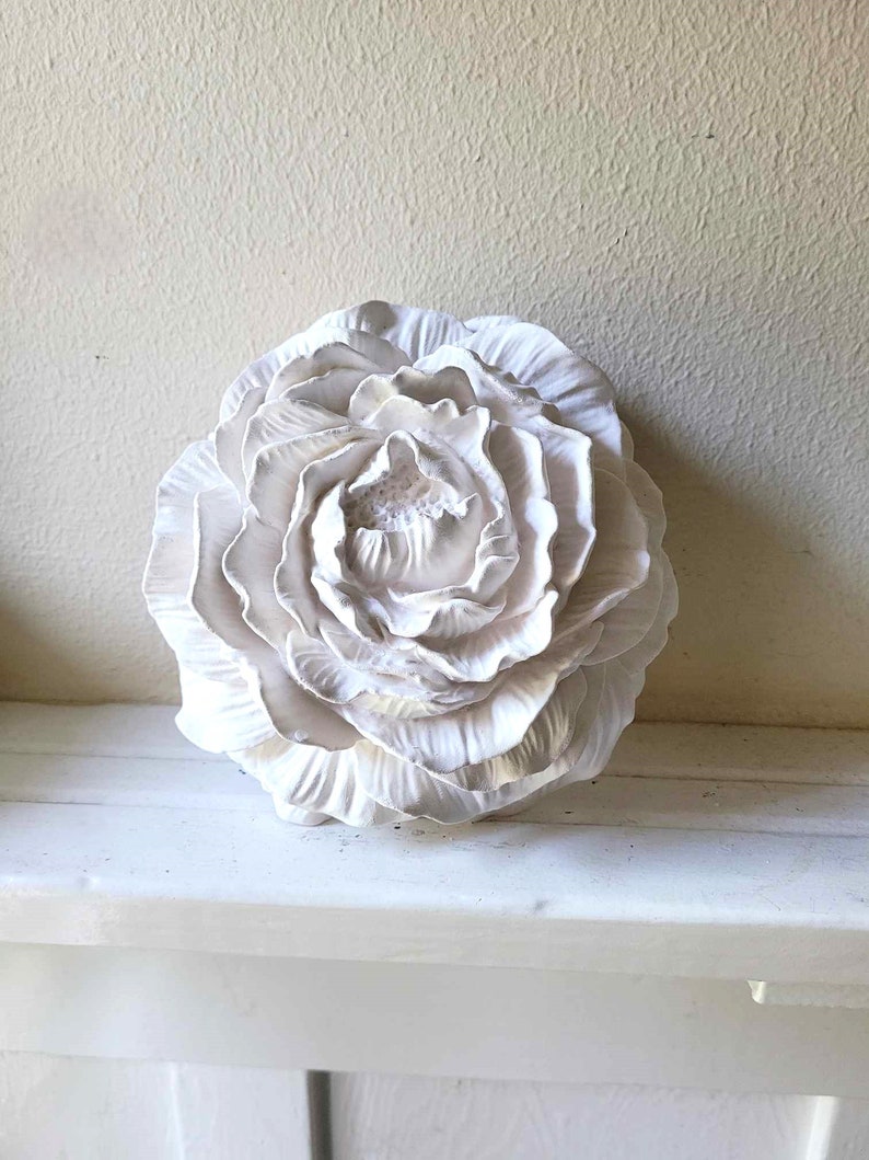 Puede incluir: Una flor de cer&aacute;mica blanca con un dise&ntilde;o detallado y en capas. La flor es una sola flor con m&uacute;ltiples p&eacute;talos.