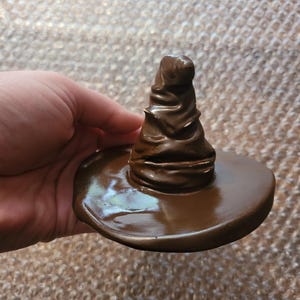 May include: A brown, miniature Sorting Hat, a replica of the iconic wizarding hat. The hat features a wide brim and a tall, pointed crown with textured details. The hat is a solid, glossy brown color.