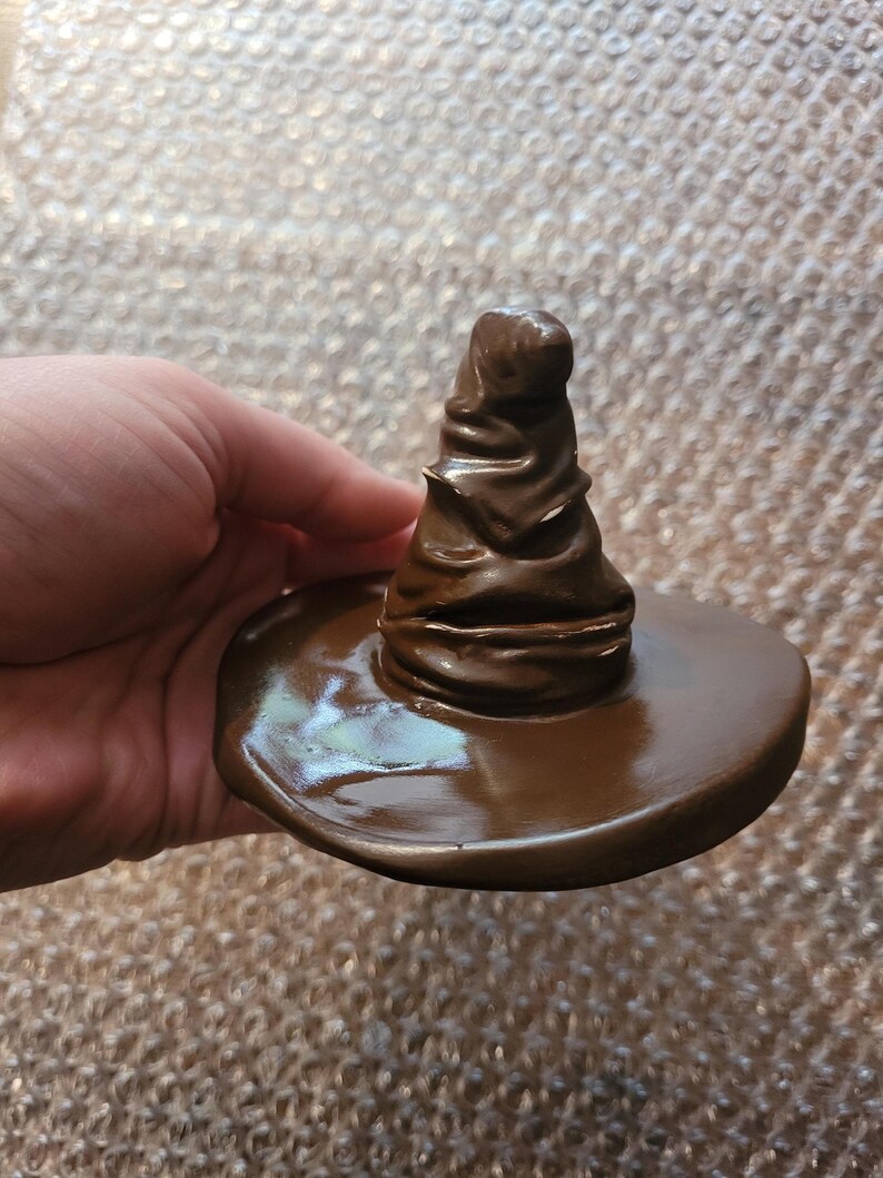 May include: A brown, miniature Sorting Hat, a replica of the iconic wizarding hat. The hat features a wide brim and a tall, pointed crown with textured details. The hat is a solid, glossy brown color.