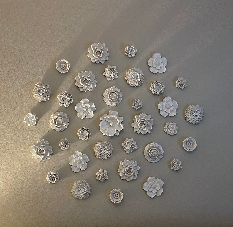 Puede incluir: Una colecci&oacute;n de decoraci&oacute;n de pared de flores de cer&aacute;mica blanca dispuestas en un patr&oacute;n circular en una pared de color beige claro. Las flores son de varios tama&ntilde;os y formas, algunas tienen una apariencia m&aacute;s realista y otras son m&aacute;s estilizadas.