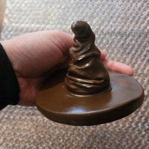 May include: A brown, glossy, decorative hat with a wide brim. The hat is a replica of the Sorting Hat from the Harry Potter series. The hat is held in a hand.