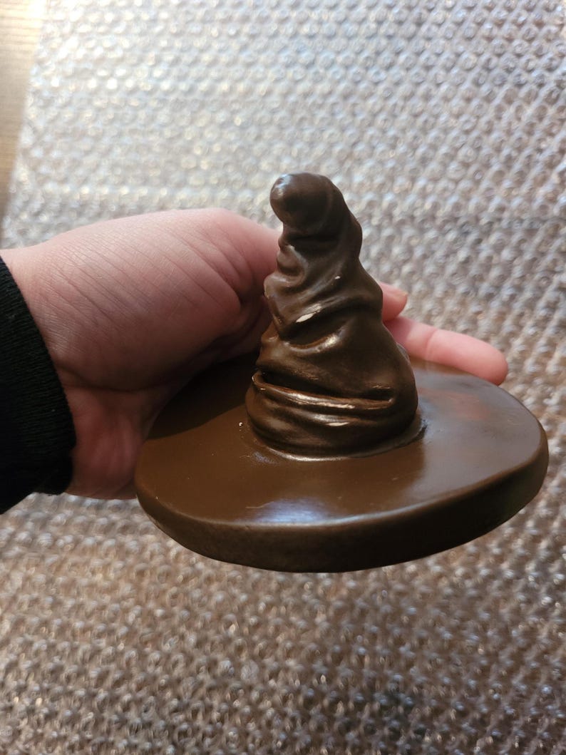 May include: A brown, glossy, decorative hat with a wide brim. The hat is a replica of the Sorting Hat from the Harry Potter series. The hat is held in a hand.