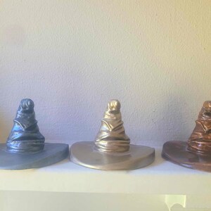 May include: Three decorative Sorting Hats in metallic finishes: silver, gold, and bronze. Each hat has a wide brim and a tall, pointed crown. The hats are displayed on a white shelf against a neutral background.