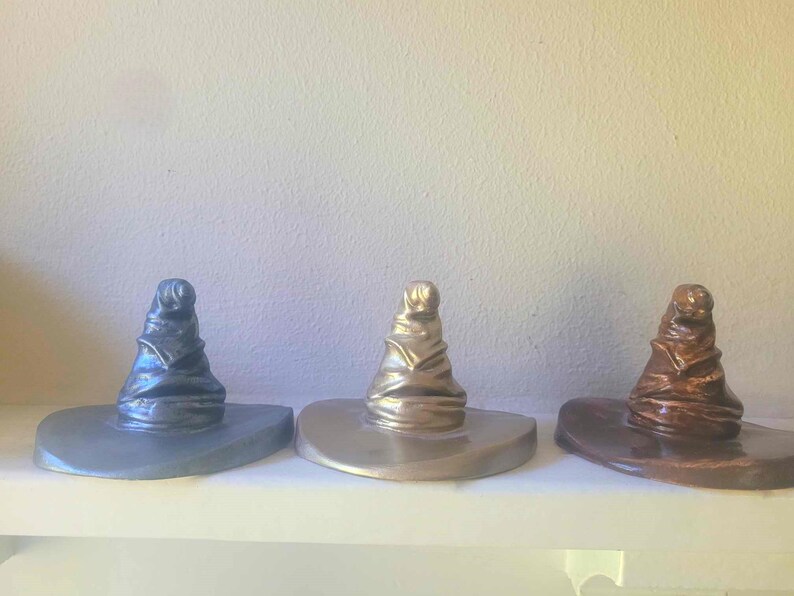 May include: Three decorative Sorting Hats in metallic finishes: silver, gold, and bronze. Each hat has a wide brim and a tall, pointed crown. The hats are displayed on a white shelf against a neutral background.