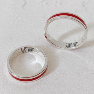 Puede incluir: Dos anillos de plata con una banda de esmalte rojo. Los anillos tienen caracteres chinos inscritos en el interior.