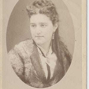 May include: A sepia-toned portrait of a woman in an oval frame. She wears a patterned jacket over a white blouse with a dark tie. Her hair is styled up with a side part, and she wears earrings. The photograph is a vintage cabinet card.