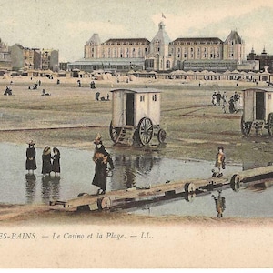 May include: A vintage postcard showing the beach at Malo-les-Bains with bathing machines, a horse-drawn carriage, and people walking on a wooden boardwalk. In the background, a large casino building stands near the sandy shore.
