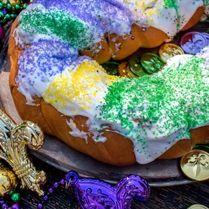 以下が含まれることがあります： 紫、緑、金色のスプリンクルで飾られた伝統的なキングケーキ。ケーキは、金と紫のマルディグラビーズとフルール・ド・リスが飾られた銀の皿の上に置かれています。