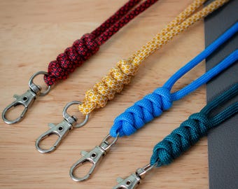 May include: Four colorful lanyards with metal swivel clips. The lanyards are red, yellow, blue, and teal, each with a woven pattern. The clips are silver. The lanyards are arranged diagonally on a light brown wooden surface.