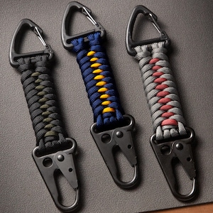 May include: Three keychains featuring braided paracord in black with olive green, navy blue with yellow, and grey with red accents. Each keychain includes a black triangular carabiner at the top and a black metal spring-loaded clip at the bottom.
