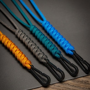 Four Paracord ID Lanyards in Goldenrod, Charcoal Grey, Teal, and Caribbean Blue, each with a black non-ferrous clip, displayed on dark leather and wood surface — handmade MRI-safe badge holders.